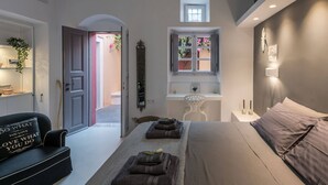 A modern bedroom with a bed, a desk, and a chair. There is a window with plants on the desk and a door leading to another room. - Coat Of Arms Mansion (Santorini)