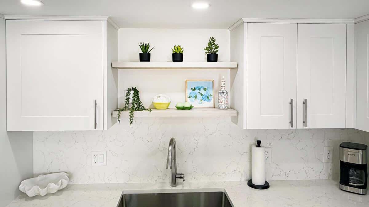 Una cocina con armarios blancos, un lavaplatos de acero inoxidable, una cafetera y plantas decorativas en estantes.