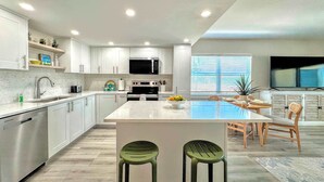 A modern kitchen with white cabinets, a central island, and a dining area with a TV. - Vacation Villas #431 Stunning Beachfront Condo (Fort Myers Beach)