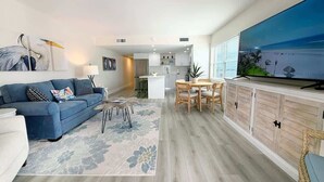 A modern living room with a sofa, coffee table, and TV. A kitchen is visible in the background. - Vacation Villas #431 Stunning Beachfront Condo (Fort Myers Beach)
