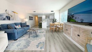 Ein modernes Wohnzimmer mit einer Couch, einem Couchtisch und einem Fernseher. Im Hintergrund ist eine Küche zu sehen.