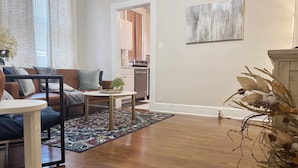 Una sala de estar con sofá, mesa de centro y alfombra. Se ve un área de cocina con armarios y encimera a través de una puerta abierta.