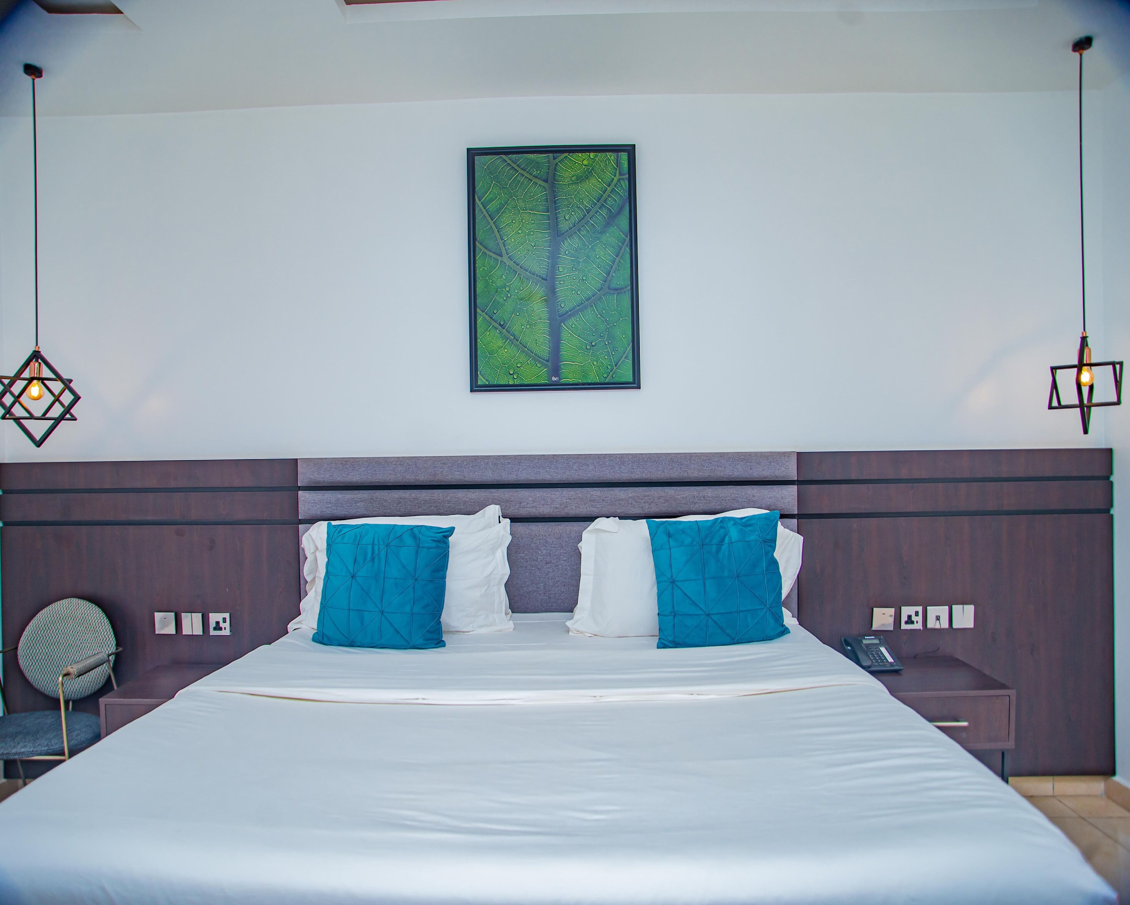 Una cama bien hecha con almohadas blancas y azules, una obra de arte con hojas enmarcada en la pared y dos lámparas colgantes.