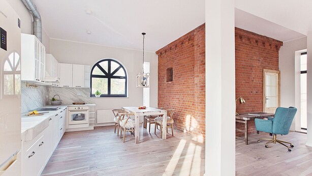 A modern kitchen with white cabinets, a large window, and a dining area with a brick wall.