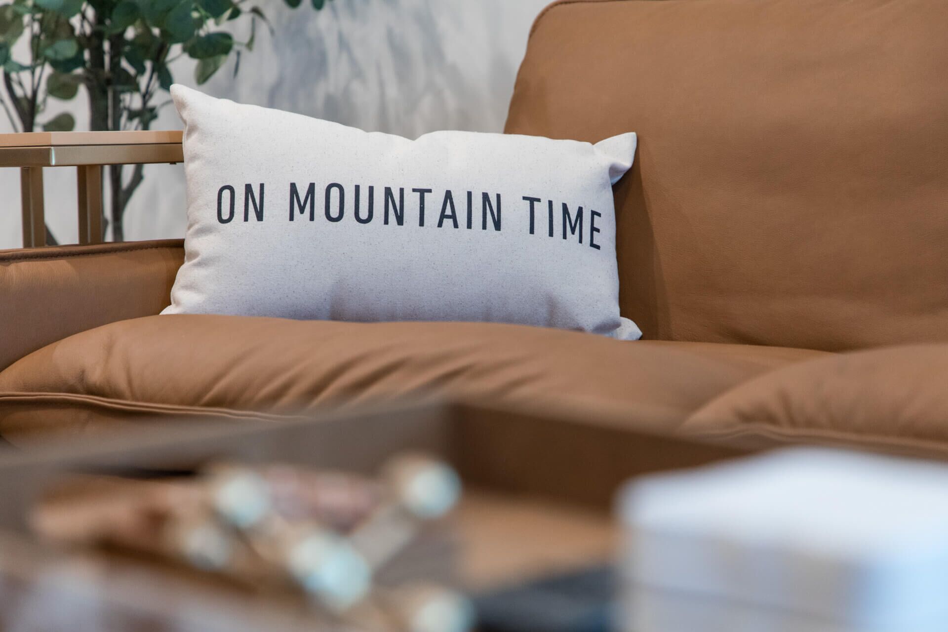 Un cuscino bianco con la scritta "ON MOUNTAIN TIME" su un divano in pelle marrone.