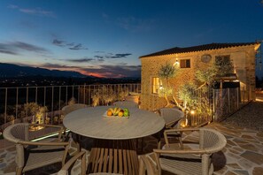 An outdoor dining area with a round table and wicker chairs, a stone building with a tiled roof, and a view of the cityscape at dusk. - Novel Villa - 5 bedrooms & Private Pool (Zakynthos)