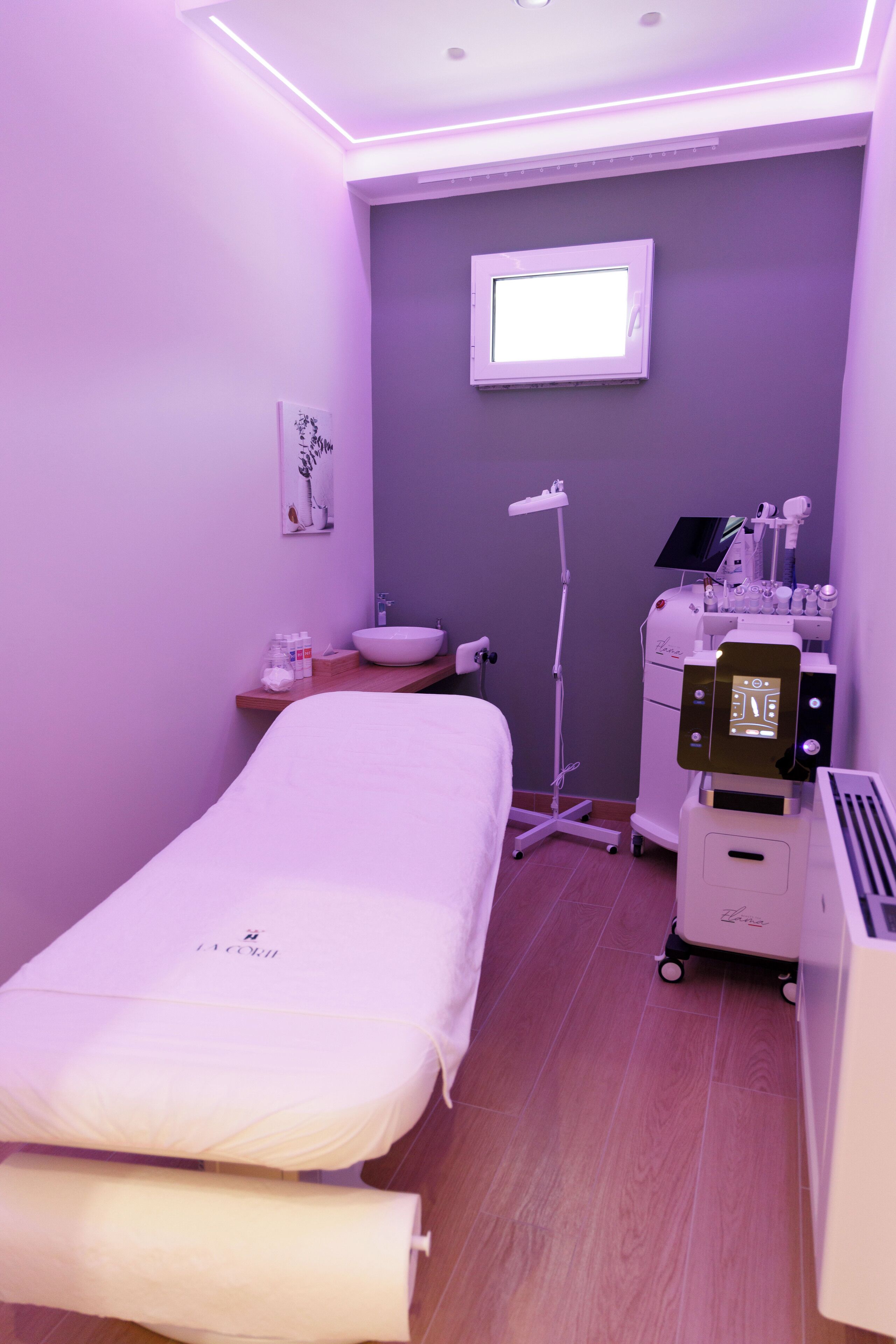 Un consultorio médico con una cama, un lavamanos, un carrito de suministros y una ventana.