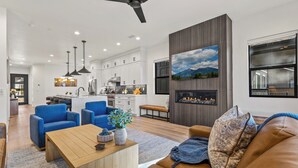 Ein modernes Wohnzimmer mit einem Couchtisch aus Holz, blauen Sesseln und einem braunen Sofa. Ein Kamin mit Fernseher darüber ist sichtbar.
