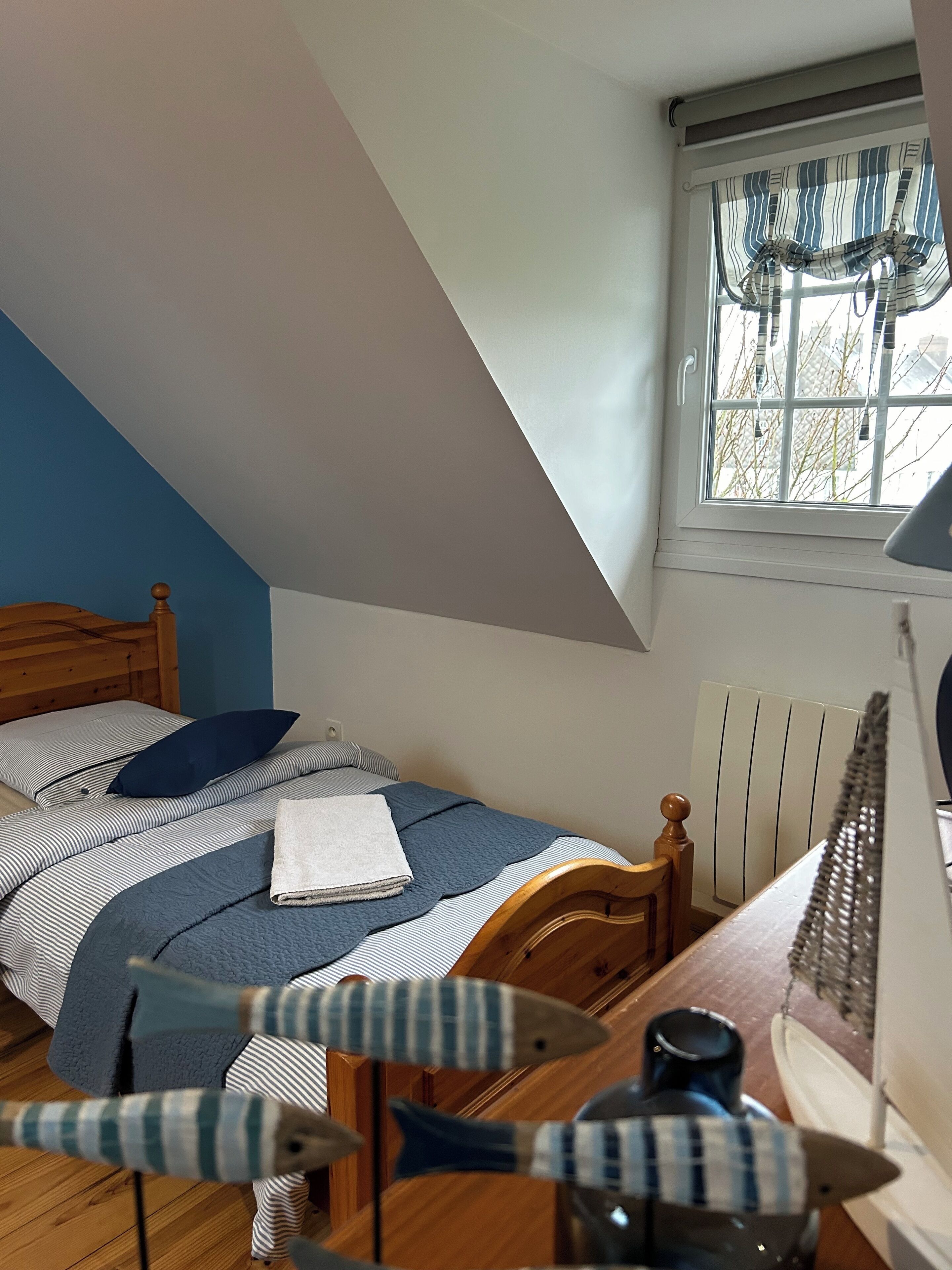 A bedroom with a sloped ceiling, a bed with striped bedding, a wooden headboard, a window with curtains, and a desk with a lamp.