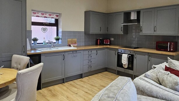 Cocina compacta con armarios grises, encimera de madera, pileta, microondas y estufa. Zona de comedor con mesa y sillas.