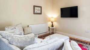 Una sala de estar con un sofá, un televisor de pantalla plana montado en la pared, un cuadro enmarcado y una lámpara sobre una mesita.