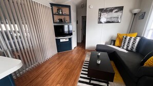 Ein modernes Wohnzimmer mit einem schwarzen Sofa, einem Couchtisch und einem Streifen Teppich. Ein Fernseher und ein Regal mit Dekorationen sind ebenfalls vorhanden.