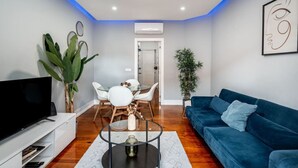 A modern living room with a blue sofa, a glass dining table with chairs, a TV on a white stand, and a large plant. - Stylish T2 Apartment in Lisbon by Lisbeyond (Lisbon)