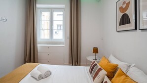 A hotel room with a bed, a nightstand, a lamp, a window with curtains, and two framed pictures on the wall. - Stylish T2 Apartment in Lisbon by Lisbeyond (Lisbon)