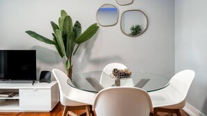 A modern dining area with a glass table surrounded by white chairs, a large potted plant, and a flat-screen TV on a white cabinet. - Stylish T2 Apartment in Lisbon by Lisbeyond (Lisbon)