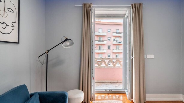 A room with a blue sofa, a floor lamp, and a view of a pink building through a window with beige curtains. - Stylish T2 Apartment in Lisbon by Lisbeyond (Lisbon)