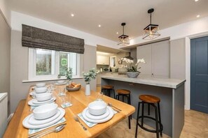 A modern kitchen with a dining area, featuring a wooden table set for a meal, a built-in island with stools, and pendant lighting. - Beck Cottage Windermere (Windermere)