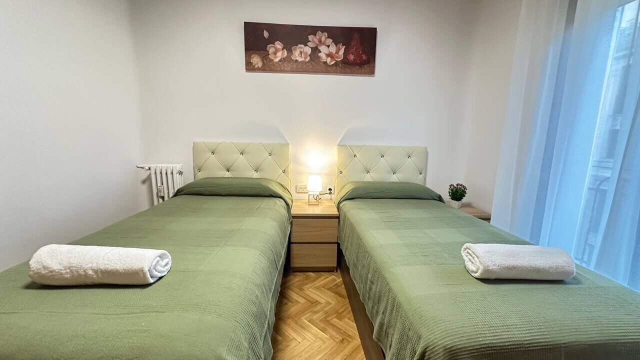 Una habitación doble con dos camas, un radiador, una mesita de noche con lámpara y un cuadro en la pared.