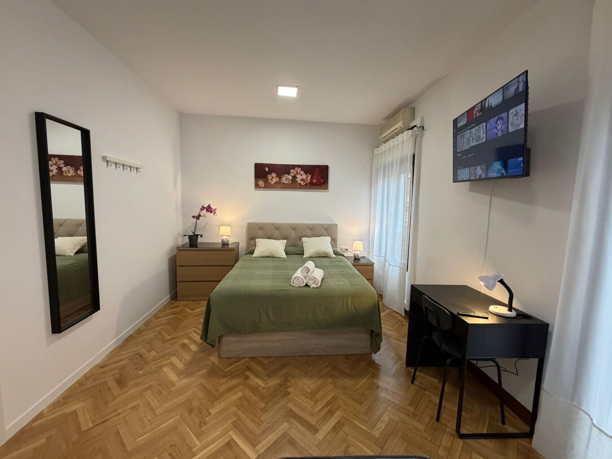 Un dormitorio con cama, escritorio, espejo, mesita de noche, planta en maceta y televisor montado en la pared.