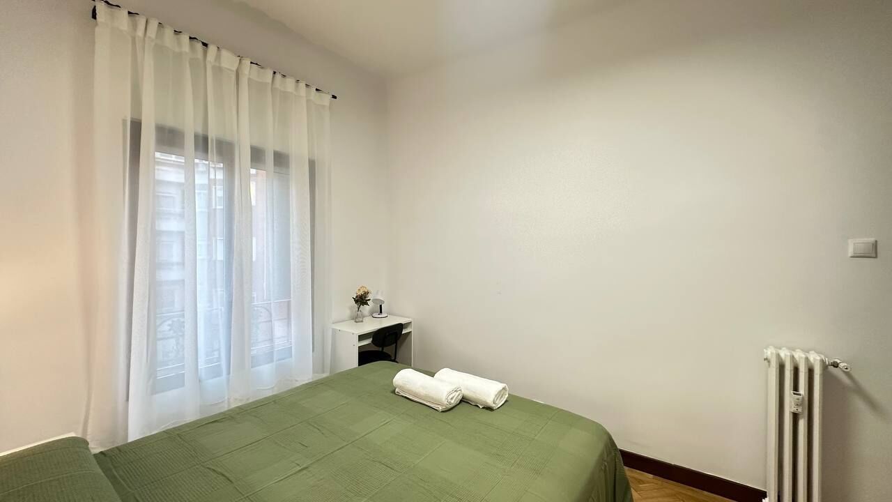 Une chambre avec un lit, un petit bureau, un radiateur et une fenêtre ornée de rideaux légers.