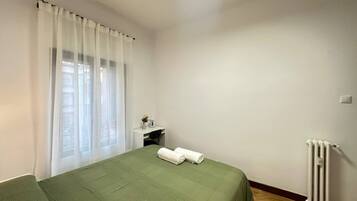 Une chambre avec un lit, un petit bureau, un radiateur et une fenêtre ornée de rideaux légers.