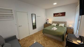 Un dormitorio con cama, mesitas de noche, escritorio, silla, televisor y puerta.