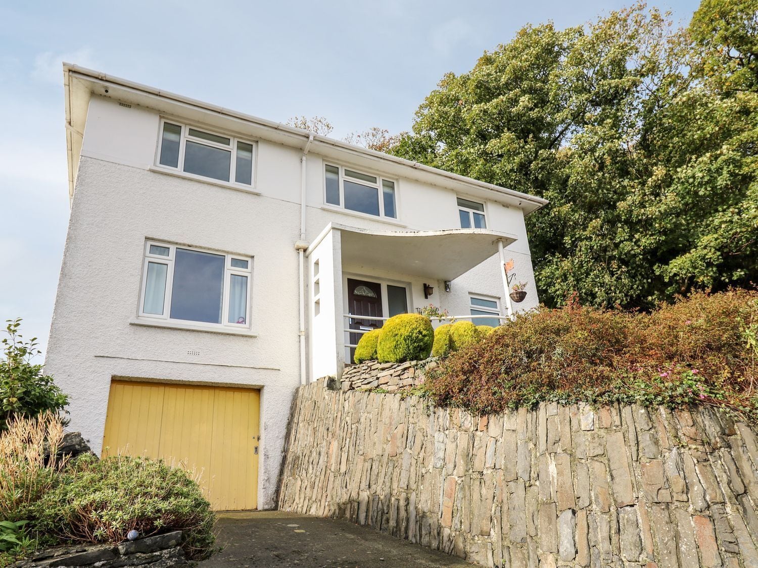 A two-story residential building with a yellow garage door, a stone wall, and a small garden area with shrubs and a potted plant.