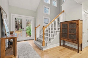 Un escalier avec des rampes blanches et des balustres en bois, menant à un palier où se trouvent un meuble en bois et un tableau encadré posé sur une table.
