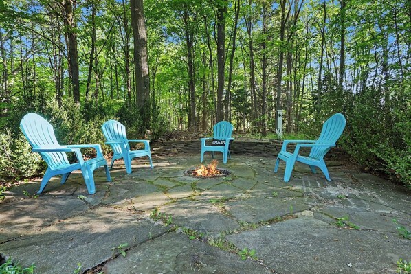 A fire pit surrounded by four Adirondack chairs in a forested area.