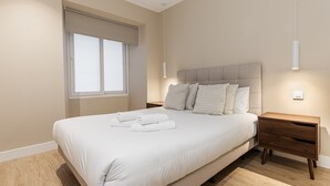 A bedroom with a large bed, a wooden nightstand, a window with blinds, and a wall-mounted light fixture. - Waou Essence VI Antón Martín - Short Duration (Madrid)