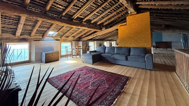 A spacious loft with exposed wooden beams, a large gray sectional sofa, a red patterned rug, and a view of the outdoors through large windows.