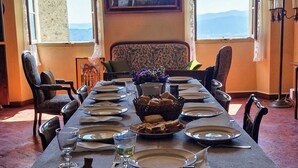 A dining room with a long table set for a meal, two windows with a view, and a chandelier hanging from the ceiling.