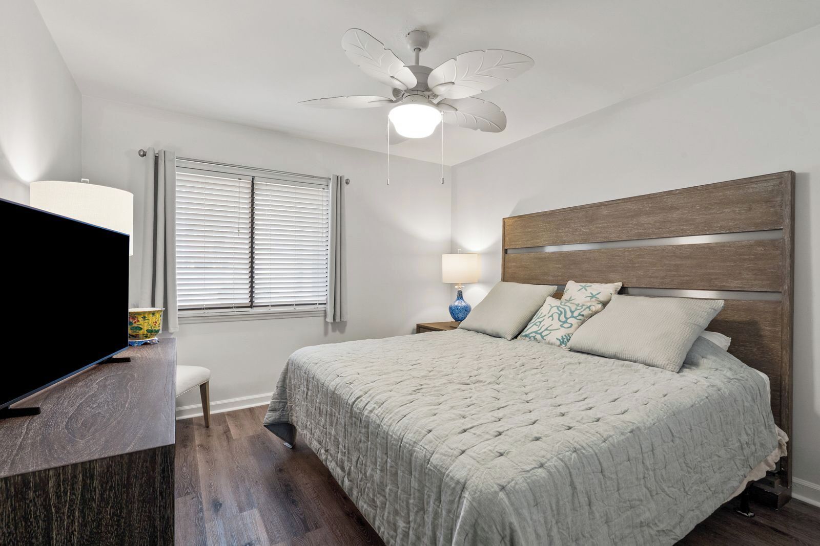 A bedroom with a bed, a ceiling fan, a TV, a lamp, and a window with blinds.