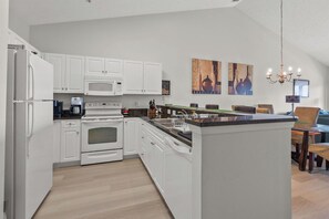 Eine moderne Küche mit weißen Schränken, einem eingebauten Ofen, einer Mikrowelle und einer zentralen Kochinsel mit Spüle. An der Wand hängen zwei Gemälde und über dem Essbereich hängt eine Kristallleuchter.