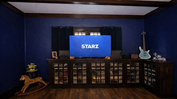 Une pièce aux murs bleu foncé, avec un plancher en bois, un téléviseur Starz posé sur un meuble, une guitare et un cheval à bascule.