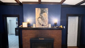 Une cheminée surmontée d’une étagère où sont exposés un tableau et des objets décoratifs, située dans une pièce aux murs bleu foncé et au plafond orné de poutres en bois.