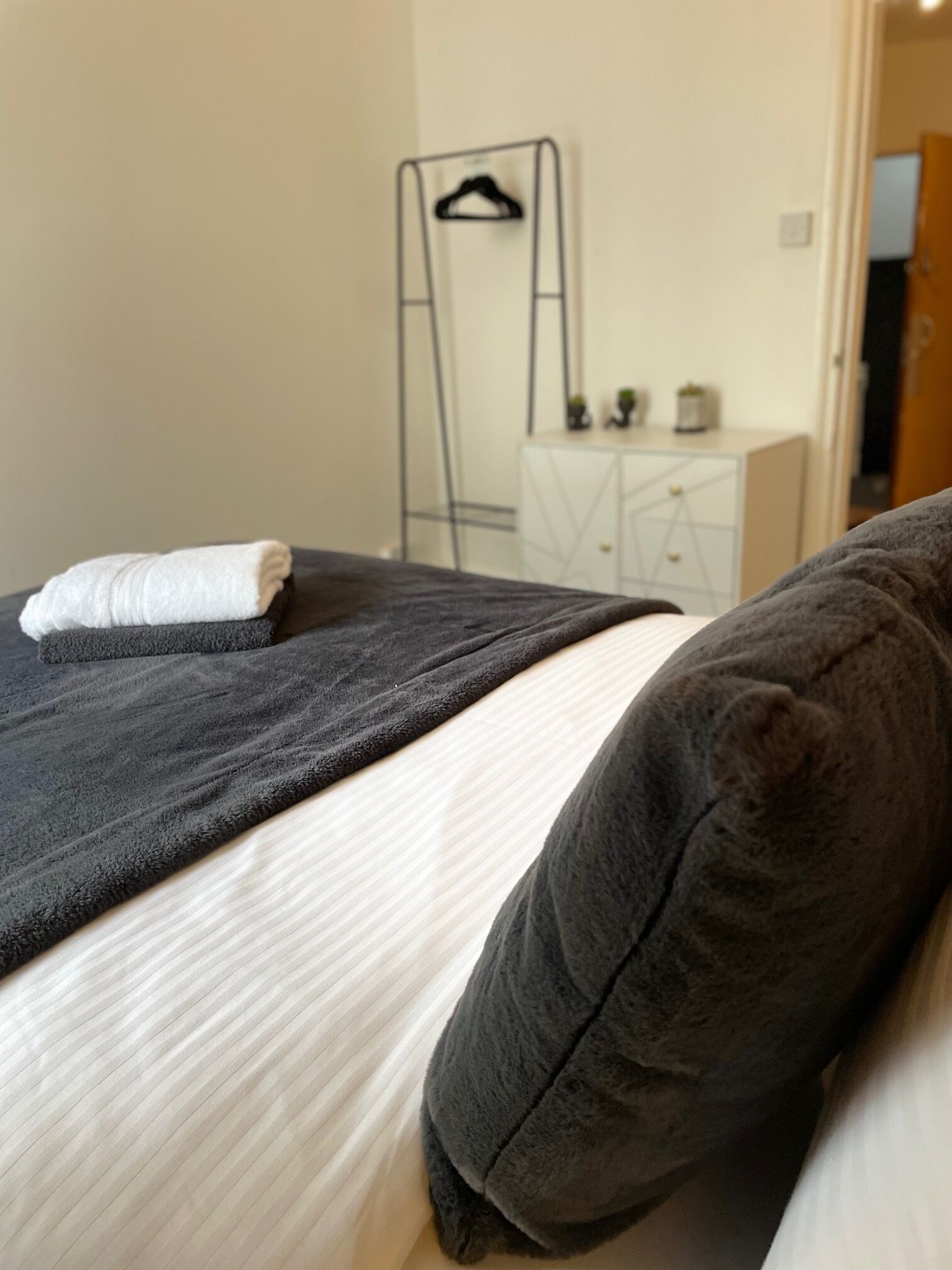A bed with a dark blanket and pillows, a white pillow, and a small white towel on the bed. There is a white dresser with a mirror and a plant in the background.
