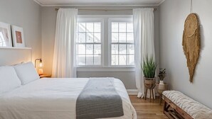 A bedroom with a large bed, a bedside lamp, a window with curtains, a wall-mounted macrame piece, and a small bench.