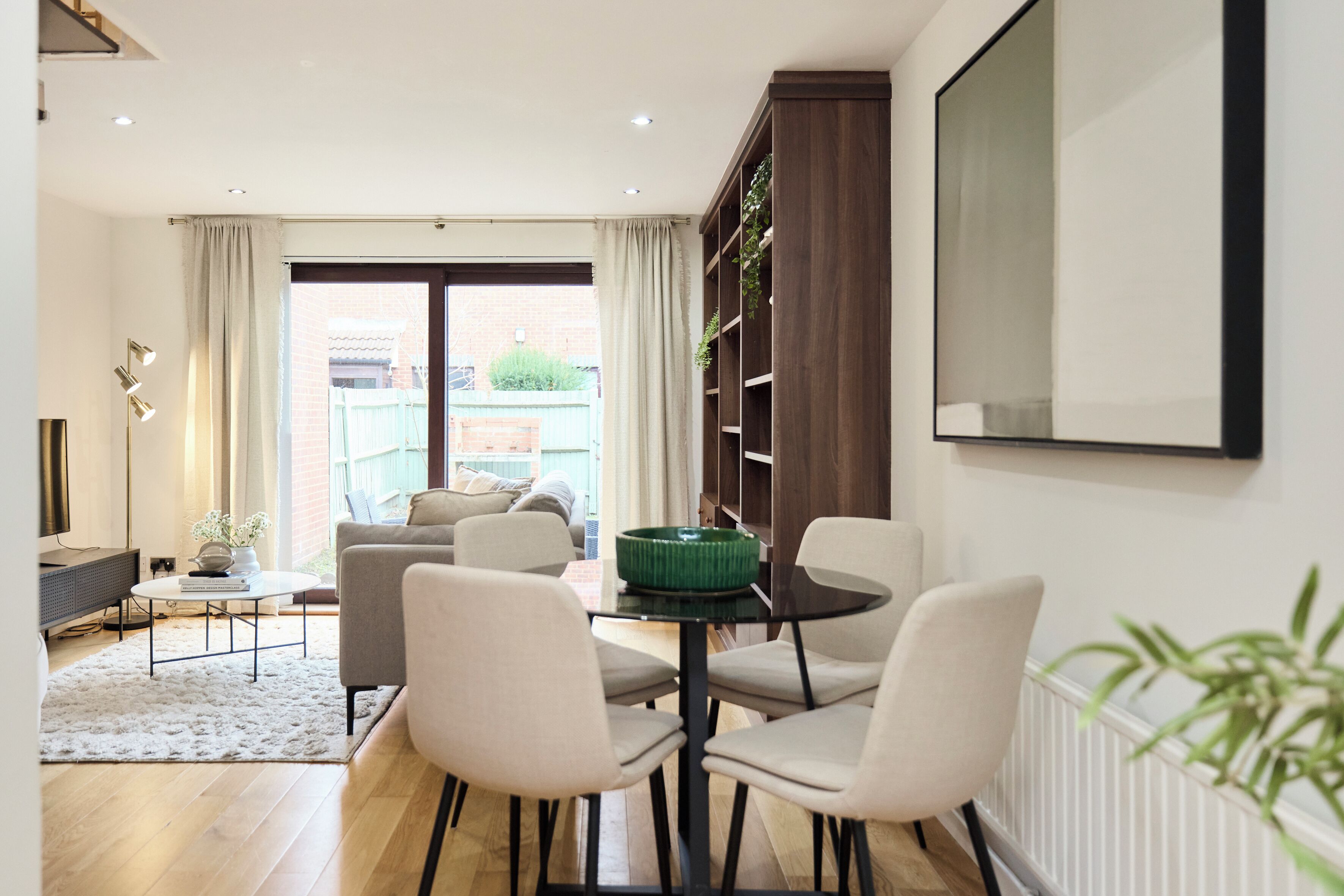 A modern living room with a sofa, chairs, a coffee table, and a dining table. There is a large window with curtains, a plant on the wall, and a mirror.
