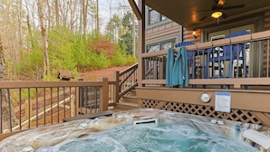 Una vasca idromassaggio su un pontile di legno con vista su alberi e un cottage in legno sullo sfondo.