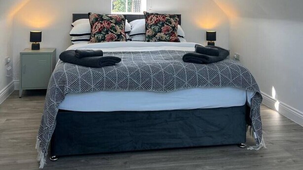 A modern bedroom with a sloped ceiling, a large bed with patterned bedding, two bedside lamps, and a window with blinds. - Gatwick 3 Bed Parking (Surrey)