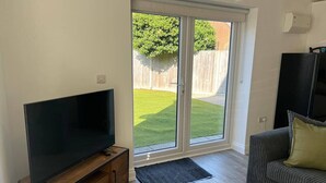 A living room with a flat-screen TV on a wooden stand, a grey sofa with a green pillow, and a sliding glass door leading to a backyard with a lawn and a wooden fence. - Gatwick 3 Bed Parking (Surrey)