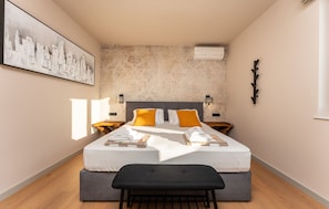 Une chambre d’hôtel moderne avec un grand lit, deux tables de chevet, un banc et un tableau représentant un paysage urbain accroché au mur.