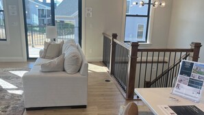 Ein geräumiges Wohnzimmer mit einem weißen Sofa, einem Esstisch mit einem Laptop und Blick durch eine Glastür nach draußen.