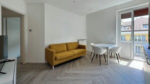 A modern living room with a mustard sofa, a white dining table, and chairs. There is a large window providing a view of the outside. - Domo House 3 (Domodossola)