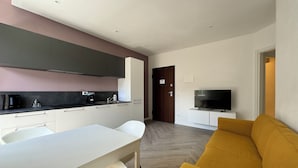A modern kitchen with white cabinets, a black countertop, and a built-in oven. There is a dining table with chairs, a sofa, and a television in the living area. - Domo House 3 (Domodossola)