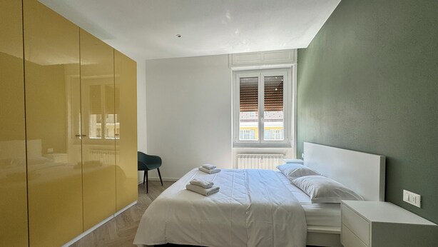 A modern bedroom with a bed, bedside table, and a window with blinds. - Domo House 3 (Domodossola)