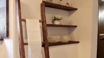 Eine Holzleiter lehnt an einer Wand, an der ein weißes Handtuch hängt. Darüber befinden sich Regale mit Körben und einer kleinen Pflanze. Ein Korbwäschekorb steht auf dem Boden neben der Leiter.