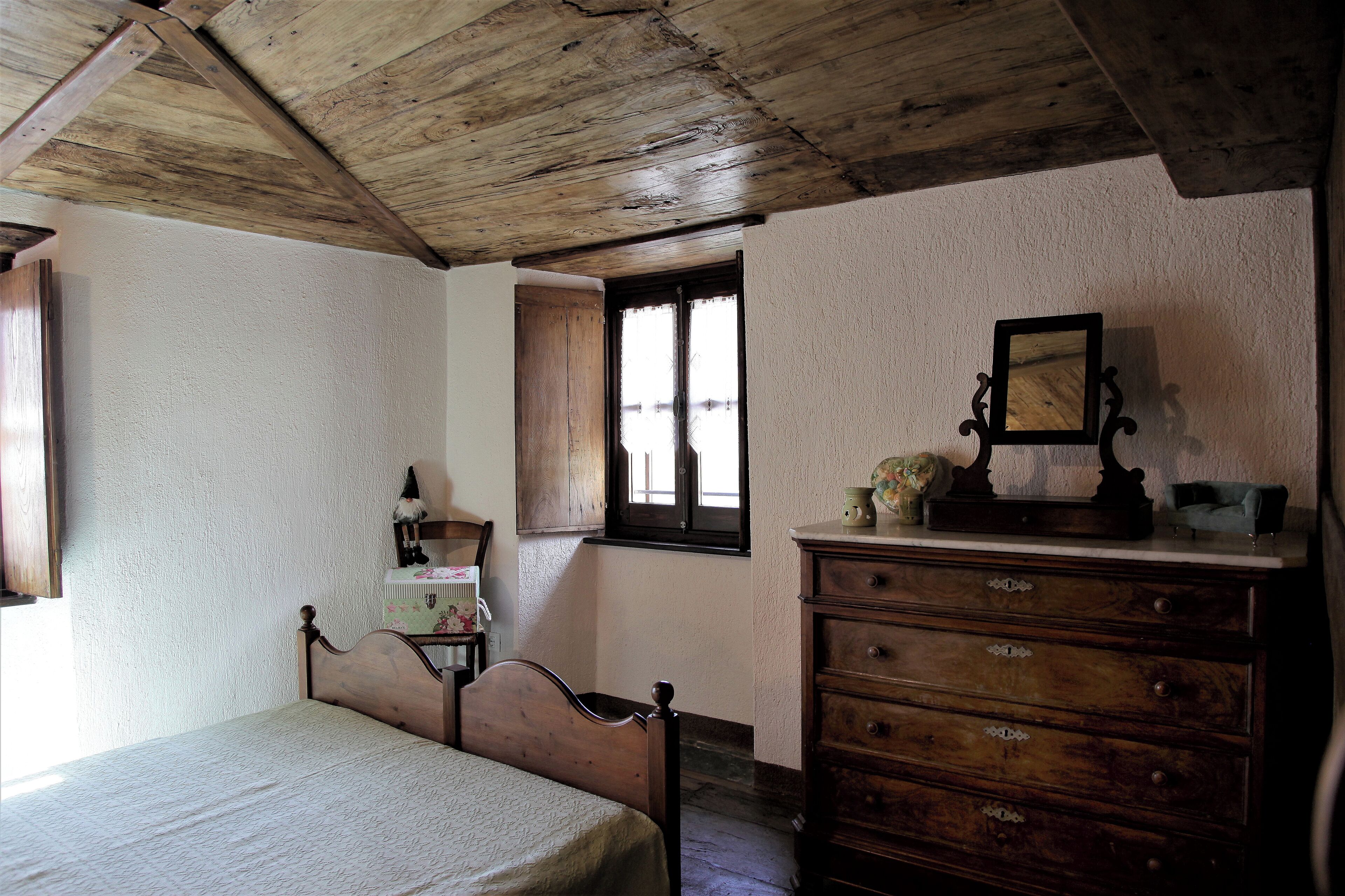 Ein Schlafzimmer mit Holzboden, einem Bett mit Holz Kopfteil, einer Kommode mit Schubladen, einem Fenster mit Holzrahmen und einem kleinen Tisch mit Lampe.