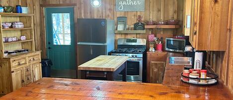 A rustic kitchen with a large wooden island, a refrigerator, stove, and a microwave. There are hanging lights and wooden shelves with kitchenware.